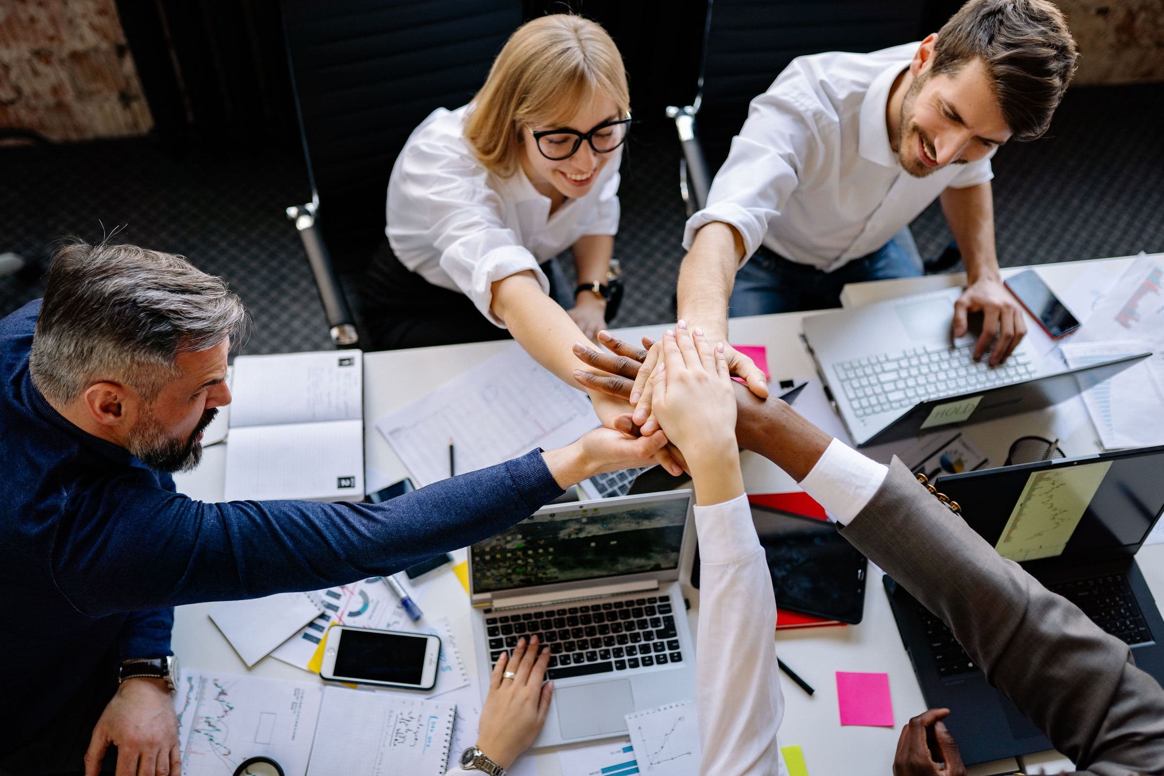 A team stacking hands together over a desk of laptops and charts
